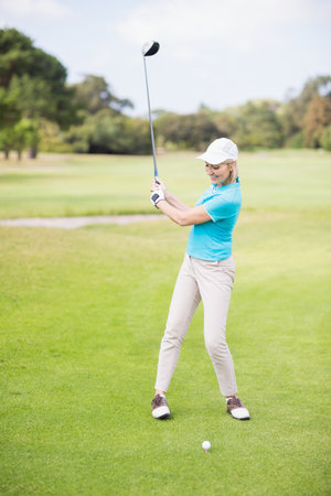 Female golfer swinging driver on golf course fairway with cap glove and golf ball on tee. Sports, recreation, elegance, outdoor, activity, leisure, athleticの写真素材