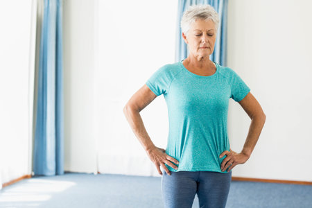 Senior female in gym clothes doing breathing exercise at home by windows with blue curtains. Fitness, wellness, serenity, mindfulness, relaxation, home environment, contemplativeの写真素材