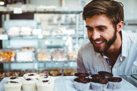 Bearded man leaning forward inspecting cupcakes behind glass case at bakery counter, copy space. Dessert, confections, sweetness, artisanal, retail, gourmet, indulgenceの写真素材