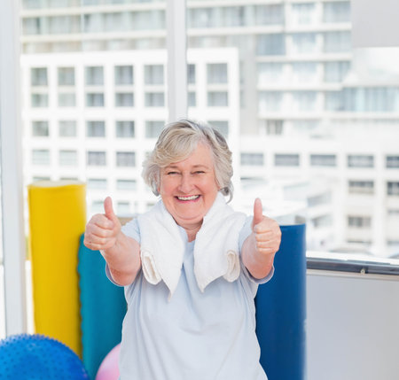 Senior woman giving thumbs up in fitness studio by window with foam rollers and exercise ball. Wellness, vitality, support, active lifestyle, modern, motivation, healthの写真素材
