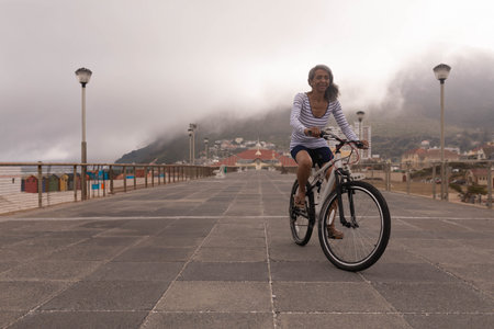 Senior woman riding bicycle along stone-paved pier near colorful beach huts under misty mountain. Scenic, leisure, vintage, coastal, nostalgic, relaxing, outdoorの写真素材