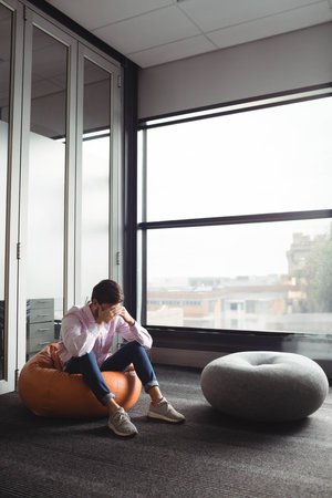 Orange beanbag chair resting beside floor-to-ceiling window in office lounge with gray pouf....