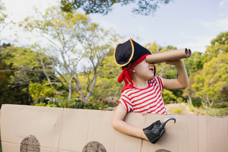 Child wearing pirate hat using cardboard ship and peering through spyglass in garden among trees. Adventure, imagination, childhood, playful, outdoor, exploration, rusticの写真素材