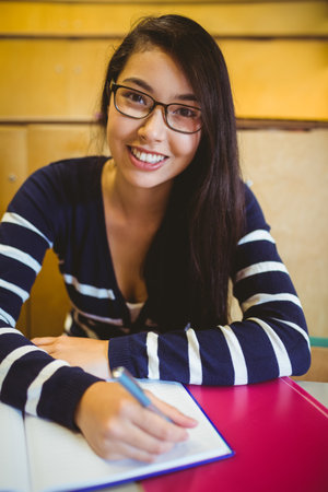 Asian female student writing notes with pen in notebook at lecture hall desk with pink binder. Education, learning, classroom, study, focus, academic, concentrationの写真素材