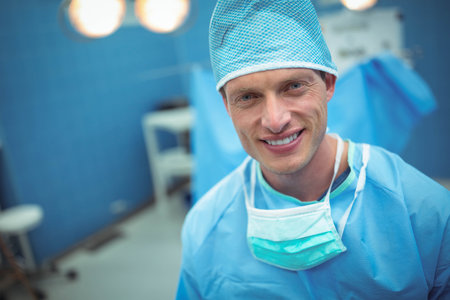 Male surgeon standing in operating room with surgical lights wearing blue scrubs, cap and mask. Medical, healthcare, professionalism, sterile, precision, clean, diagnosticの写真素材