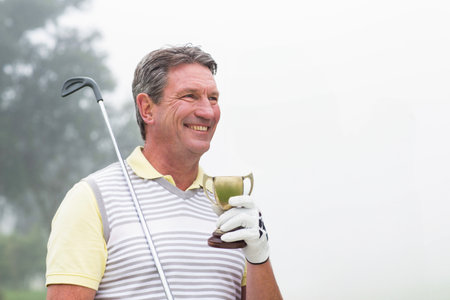 Senior man in golf attire holding trophy and club on misty fairway, copy space. Luxury, achievement,...