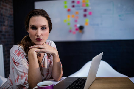 Woman in late twenties leaning over laptop at office with whiteboard with sticky notes, copy space. Collaboration, brainstorming, workspace, modern, productivity, team, communicationの写真素材