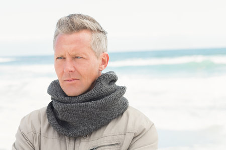 Man in early 50s standing on sandy beach wearing beige zip-up jacket and grey knit scarf. Landscape, maturity, contemplative, coastal, outdoor, thoughtful, apparelの写真素材