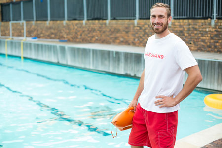 Male lifeguard smiling while holding orange rescue buoy on pool deck near lane ropes, copy space. Rescue, safety, outdoor, aquatic, vigilant, modern, leisureの写真素材