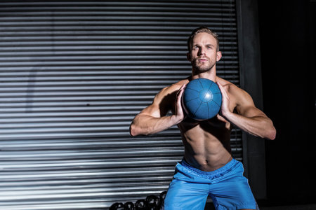 Shirtless athletic man holding medicine ball and squatting beside kettlebells at gym, copy space. Strength, training, fitness, muscular, workout, strength training, sportsの写真素材