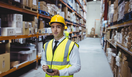 Male warehouse worker wearing hardhat reflective vest in aisle scanning boxes on racks with scanner....