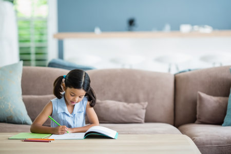 Child writing in open notebook with colored pencils at wood coffee table in living room. Creativity,...