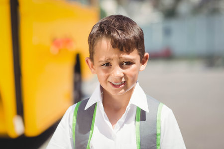 Asian child standing and waiting by yellow school bus on sunny pickup area with grey-green backpack....