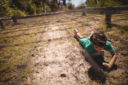 Boy is crawling under nylon obstacle net on sandy path casting grid shadows in wooded park. Adventure, outdoor, play, exploration, activity, natural, dynamicの写真素材