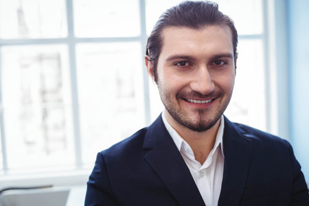 Man in early thirties wearing business suit standing and smiling in office near multi-pane window....
