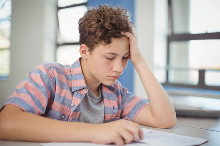 Teenage boy in striped shirt leaning forward and writing on paper at school desk with notebook....
