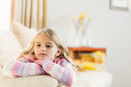 Little girl in knitted sweater leaning on sofa at home near vase with flowers, copy space. Cozy, serenity, pastel, relaxation, interior, warmth, innocenceの写真素材