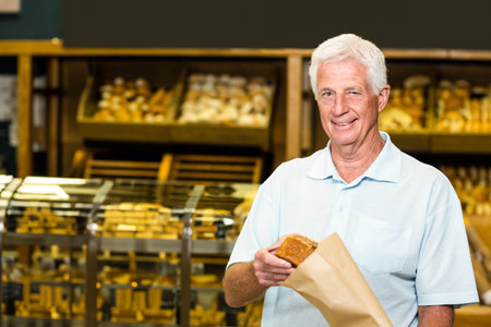 Senior man smiling and holding loaf of bread in paper bag inside bakery shop, copy space. Artisan, heritage, rustic, warmth, leisure, craftsmanship, comfortの写真素材