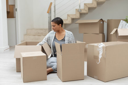 African American woman sitting in foyer by staircase rail unpacking cardboard boxes and bubble wrap. Settling, organizing, home, transition, decor, renovation, lifestyleの写真素材