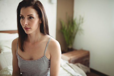Woman sitting on edge of bed wearing striped tank top in bedroom near bench holding plant. Relaxation, serenity, minimalism, cozy, natural light, interior, lifestyleの写真素材