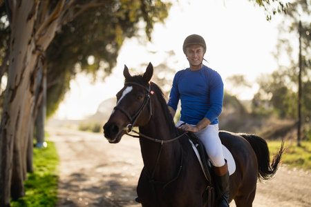 Man riding horse on sunlit trail flanked by eucalyptus trees wearing helmet and riding boots. Equestrian, outdoor, adventure, natural, wilderness, harmony, countryside