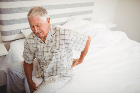 Man wearing plaid shirt and striped pants sitting on bed edge, grasping lower back, copy space. Senior, wellness, relaxation, healthcare, comfort, home, lifestyleの写真素材