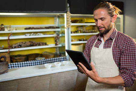 Male baker holding tablet sorting loaves on checkered cloth at bakery counter, copy space. Artisan,...
