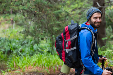 Man in late twenties walking forest trail with red backpack and hiking poles, copy space. Adventure,...