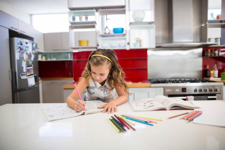 Girl child sitting at white countertop island in kitchen coloring workbook with colored pencils. Playful, learning, creativity, contemporary, home, education, vibrantの写真素材