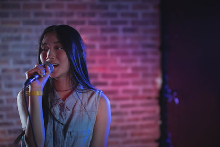Asian woman singing into microphone on stand on club stage wearing sleeveless denim top, copy space. Performance, musician, entertainer, microphone, concert, stage, ambianceの写真素材