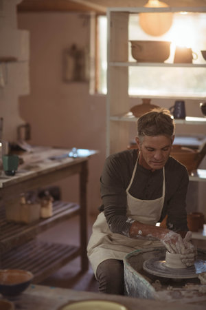 Male potter shaping clay on pottery wheel in workshop with wooden shelves and glaze containers. Craftsmanship, artisan, ceramics, handmade, studio, creative, designの写真素材