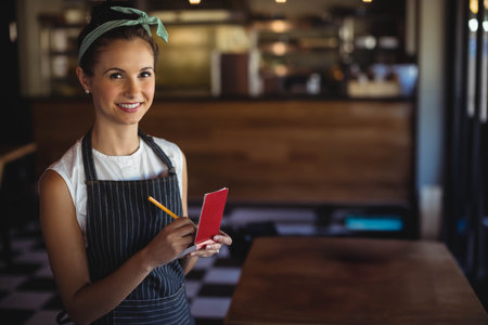 Smiling female server wearing apron at cafe table holding red order pad and pencil, copy space....