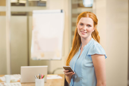 Professional woman holding smartphone while standing near office desk with laptop and flipchart....