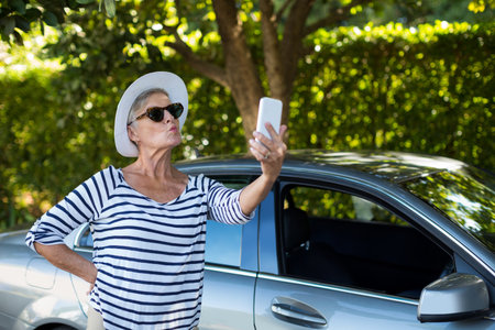 Senior woman in sun hat and sunglasses taking selfie on driveway with smartphone near silver sedan. Lifestyle, outdoor, leisure, fashion, mobile, tranquility, natureの写真素材