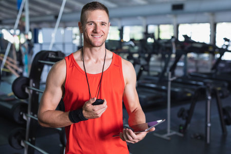 Mid adult trainer wearing athletic top holding stopwatch tablet in gym near weight benches, mirrors. Fitness, training, motivation, health, exercise, intensity, wellnessの写真素材