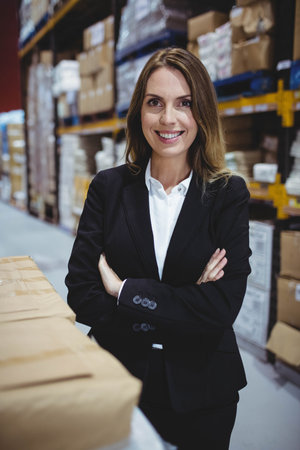 Middle-aged woman in business attire standing confidently in warehouse center among cardboard boxes. Professional, industrial, minimalist, logistics, organizational, confident, corporateの写真素材