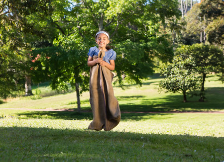 Girl jumping through burlap sack on lawn in sunshine wearing light blue top and white headband. Child, outdoor recreation, playful, vibrant, nature, activity, happinessの写真素材