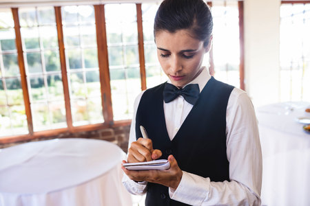 Female server in uniform writing on notepad at banquet hall by white tables, glassware, copy space. Hospitality, event, interior, decor, elegant, professional, diningの写真素材