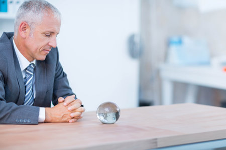 Senior businessman wearing suit sitting behind desk focusing on crystal ball in office, copy space. Professional, executive, modern, corporate, contemplative, sophisticated, minimalistの写真素材
