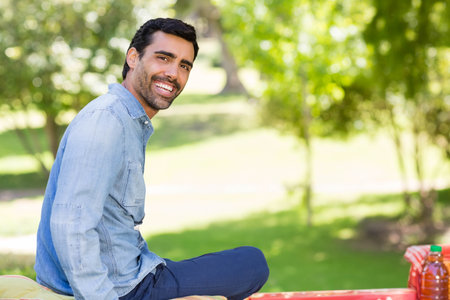 Mid adult man sitting on red picnic bench smiling in park with juice bottles, copy space. Outdoor, leisure, relaxation, nature, wellness, casual, lifestyleの写真素材