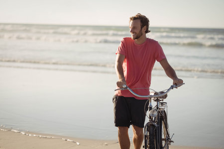 Man walking along wet sand at beach edge while pushing black cruiser bicycle, copy space. Coastline, leisure, minimalist, adventure, outdoor, tranquility, lifestyleの写真素材