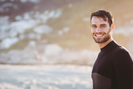 Adult man standing on sandy shore wearing wetsuit smiling at camera under golden light, copy space. Adventure, leisure, ocean, resilience, outdoor, vibrant, pastimeの写真素材