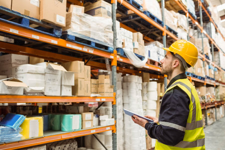 Man inspecting boxes on shelving in aisle wearing safety helmet and vest holding tablet, copy space....