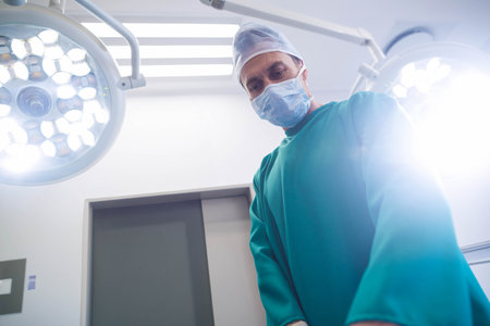 Male surgeon standing in scrubs under lamps in operating room, inspecting surgical field. Medical, healthcare, hospital, precision, sterile, professional, clinicalの写真素材