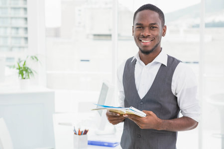 African American man standing and holding notebook in bright office with monitor, plant, copy space. Professional, workspace, modern, corporate, entrepreneurship, confident, productivityの写真素材
