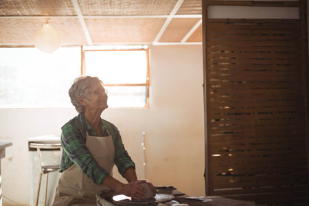 Senior woman shaping clay on pottery wheel in lit workshop wearing apron with window and lamp. Ceramics, craftsmanship, artisanal, handmade, creative, rustic, hobbyの写真素材