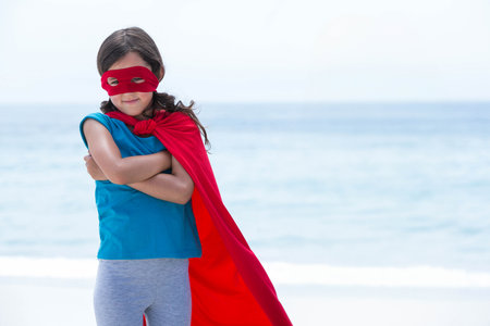 Female child striking superhero pose and standing on sandy shore wearing red cape and mask. Adventure, empowerment, youthful, heroism, confidence, outdoor, fantasyの写真素材