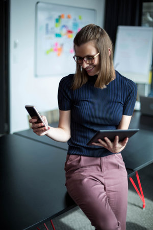 Woman leaning on conference table holding devices with whiteboard, notes and flipchart at office....