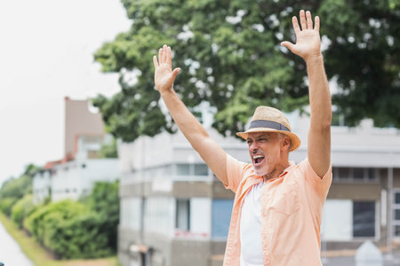 Senior man raising arms shouting beside canal path wearing straw fedora hat peach shirt, copy space. Outdoor, leisure, lifestyle, modern, wellness, nature, celebrationの写真素材
