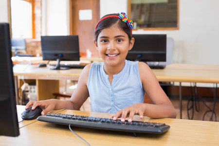 Indian schoolgirl wearing headband smiling at monitor in computer lab using keyboard and mouse....
